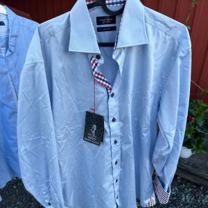 Ljusblå skjorta från Ernst Alexis - Säljer en stilren ljusblå skjorta från Ernst Alexis. Skjortan har långa ärmar och en snygg rutig detalj i kragen och längs knappslån i rött, vitt och blått. Perfekt för både vardag och fest! Aldrig använd med lappar kvar.