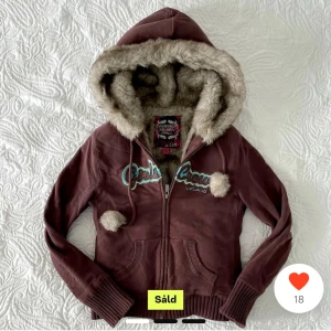 Brun hoodie med pälsfodrad luva - Mysig brun hoodie från Gale Crew med pälsfodrad luva och dragkedja. Den har en broderad logga i blått och vitt på framsidan samt dekorativa tofsar på snörena. Perfekt för kyliga dagar. Inga deffekter. Säljer då armarna är för korta för mig