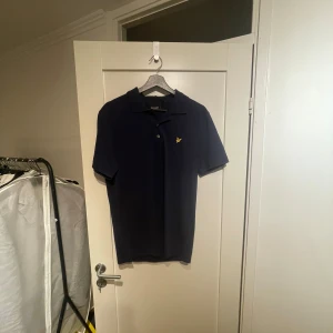 Mörkblå pikétröja från Lyle & Scott - Säljer en stilren mörkblå pikétröja från Lyle & Scott med klassisk krage och knappslå. Tröjan har en liten gul logotyp på bröstet som ger en snygg kontrast. Perfekt för en avslappnad men ändå uppklädd look.