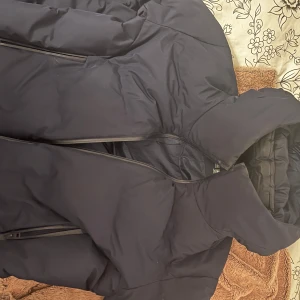 Svart dunjacka från Uniqlo - Snygg och varm svart dunjacka från Uniqlo med dragkedja och huva. Perfekt för kyliga dagar med sin quiltade design som håller värmen inne. Jackan har en stilren look och praktiska fickor.