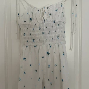 Vit blommig romper med smala axelband - Somrig vit romper (överdel med shorts) med blå blommor och smockdetaljer. Den har smala axelband och knytning framtill som kan knytas i rosetter. Alla pris kan diskuteras!