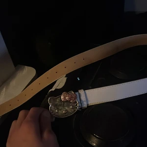 Vitt bälte med Hello Kitty-spänne - Snyggt vitt bälte med ett stort Hello Kitty-spänne i silver och en glittrig rosa rosett. Perfekt för att ge en lekfull touch till din outfit. Bältet har flera hål för justerbar passform. 105 cm långt