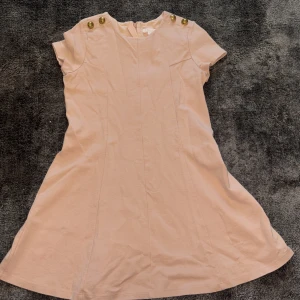 Beige klänning från Chloé - Elegant rosa beige klänning från Chloé med korta ärmar och dekorativa guldknappar på axlarna. Perfekt för en stilren look.