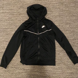 Svart hoodie från Nike - Snygg svart hoodie från Nike storlek s/xs med dragkedja och vit logga på bröstet. Perfekt för en sportig look eller avslappnade dagar. Den har en huva och långa ärmar för extra komfort. Skick 8-10 pris 400