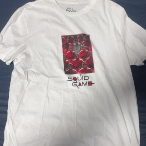 Vit Squid Game t-shirt - Snygg vit t-shirt med tryck från Squid Game. Designen visar flera karaktärer i rött med texten 'Squid Game' under. Perfekt för fans av serien!