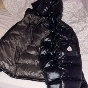 Svart dunjacka från Moncler - Snygg svart dunjacka från Moncler med glansig finish och huva. Jackan har en dragkedja och är perfekt för kyliga dagar. Den ikoniska Moncler-loggan syns på ärmen.
