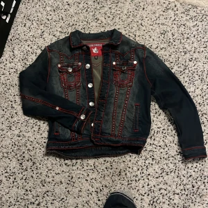 Jeansjacka från True Religion - Snygg jeansjacka från True Religion med röda sömmar och silverfärgade knappar. Jackan har två bröstfickor med lock och knappar samt en klassisk krage. Perfekt för en cool och avslappnad stil.