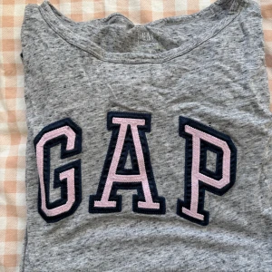 Grå blouse från GAP - Säljer en grå t-shirt från GAP med stort rosa och svart logotryck på framsidan. Perfekt för en avslappnad stil.