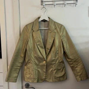 Guldglänsande kavaj - Snygg guldglänsande kavaj med klassisk krage och en knapp framtill. Ser större ut en vad den är på bilderna. Passar mig som är en 36/38. Slutar över rumpan och är hyfsat tajt i midjan. Verkligen super snygg men har tyvärr inte kommit till användning 