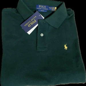 Helt ny. Mörkgrön pikétröja från Ralph Lauren - Snygg mörkgrön pikétröja från Ralph Lauren med klassisk krage och knappslå. Den har det ikoniska broderade logotypen i gult på bröstet. Perfekt för en stilren look. Storlek 14-16