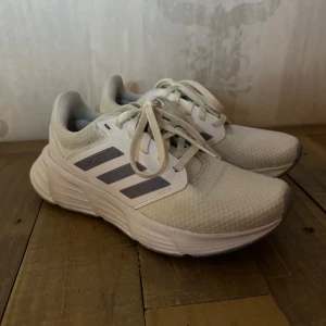 Vita sneakers från Adidas - Säljer ett par vita sneakers från Adidas med klassiska tre ränder i grått. Skorna har snörning och en bekväm sula, perfekt både till idrotten & vardags! Skorna är inte mycket använda, endast några gånger inomhus. Jättefina!