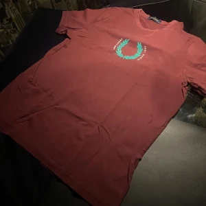 Röd t-shirt från Fred Perry - Snygg röd t-shirt från Fred Perry med ett grönt lagerkransmotiv och texten 'Since 1952'. Perfekt för en avslappnad stil.