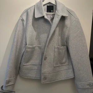 Grå kappa från 157 - Snygg grå kappa från 157 med stora fickor och knappar framtill. Perfekt för kyligare dagar med sin stilrena design och bekväma passform.