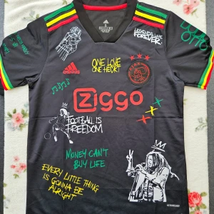 Svart fotbollströja med Bob Marley-tema - Snygg svart fotbollströja från Adidas med Bob Marley-tema. Tröjan har korta ärmar och färgglada detaljer i rött, gult och grönt. Den har tryck som 'One Love One Heart' och 'Football is Freedom'. Perfekt för fotbollsfans som älskar reggae och Bob Marley.