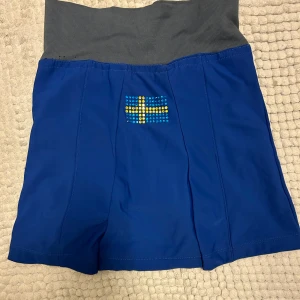 Blå träningskjol med svenska flaggan - Träningskjol, blå, med svensk flagga i glittriga stenar på baksidan. Kjolen har en bred, grå resår och inbyggda shorts för extra komfort. Perfekt för sport och fritid!