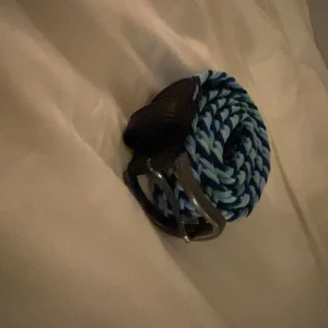 Flätat bälte i blå nyanser 110 cm långt - Snyggt flätat bälte i olika blå nyanser med ett stilrent silverfärgat spänne. Bältet har detaljer i mörkt skinn vid ändarna som ger en elegant touch. Perfekt för att liva upp vilken outfit som helst!