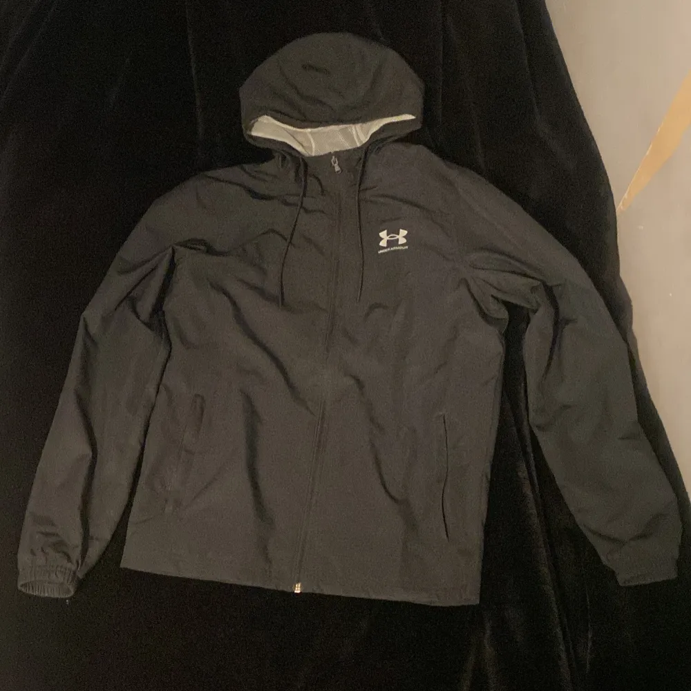 Säljer en svart vindjacka från Under Armour med huva och dragkedja. Jackan har en lös passform och är perfekt för blåsiga dagar. Den är tillverkad i syntetmaterial och har ett meshfoder för extra komfort. Vid minsta lilla intresse är det bara att skriva. Takit.