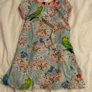 Blommig klänning med fågelmönster från Molo - Charmig klänning från Molo med ett färgglatt mönster av fåglar och blommor på en ljusblå bakgrund. Klänningen har korta ärmar med rosa kant och en rund halsringning. Perfekt för en lekfull stil! Denna fina klänning kommer tvättas och strykas innan den skickas! ⭐️