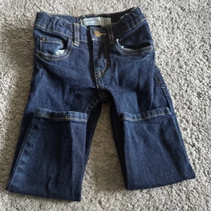 Mörkblå jeansbyxor för barn - Säljer ett par mörkblå jeansbyxor för barn med klassisk femficksdesign och knappstängning. Byxorna har en rak passform och är perfekta för vardagsbruk.