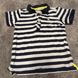 Randig pikétröja med krage - Snygg randig pikétröja i marinblått och vitt med korta ärmar och krage. Tröjan har knappar framtill och en liten broderad detalj på bröstet. Perfekt för en avslappnad stil.