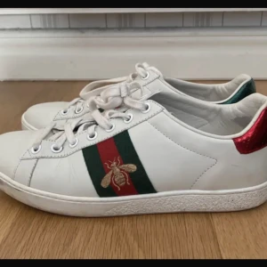 Vita sneakers med bi-motiv från Gucci - ÄKTA Gucci Ace sneakers, användt men ok skick. Storlek 36 men passar mig som normalt har 37 Ett skosnöre är trasigt men går att fixa nya, har bara knutit ihop dom nu på sidan.  smått creasade enligt bild.Kvitto finns. . Nypris ca 6900kr
