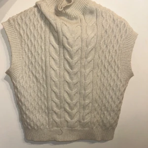 Beige stickad väst, Sthlm still  - Snygg beige stickad väst med flätmönster och hög krage. Perfekt för lager-på-lager under kyliga dagar.