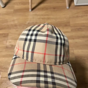 Rutig keps från Burberry - Snygg rutig keps från Burberry i klassiskt beige, svart och rött mönster. Kepsen har en broderad logotyp på framsidan och justerbar passform. Perfekt för en stilren look.
