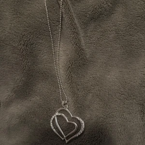 Halsband med dubbla hjärtan i silver - Elegant halsband med två sammanflätade hjärtan i silver. Det yttre hjärtat är dekorerat med glittrande stenar som ger en lyxig känsla. Perfekt för att ge en romantisk touch till din outfit.