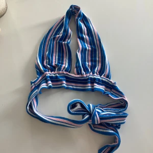 Randig bikiniöverdel i blått och rosa - Säljer en superfin topp med randigt mönster i blått, rosa och vitt. Den har en knytning i ryggen och är perfekt för sommarens alla äventyr. Materialet känns mjukt och stretchigt, vilket ger en bekväm passform.