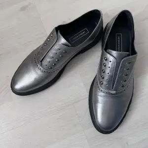 Snygga och stilrena silvriga läder loafers. Skorna har en cool design med nitar på ovansidan och en bekväm passform. Perfekta för att ge din outfit en extra edge!