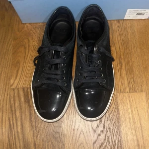 Svarta sneakers från Lanvin - Säljer ett par stilrena blå  sneakers från Lanvin. De har en snygg glansig finish på tån och en vit sula som ger en cool kontrast. Skorna har klassisk snörning och är perfekta för både vardag . De är i bra skick 9/10. Är blå men ser ut som svarta 
