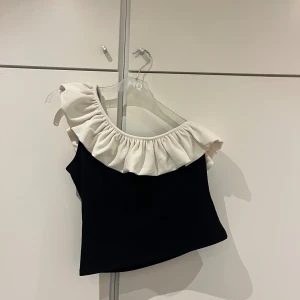 Oneshoulder topp med vit volang, HM - Volangtopp i svart med vit volang. Är oneshoulder.  Strl M. Använd 1 gång, tvättad 1 gång. Nyskick. 
