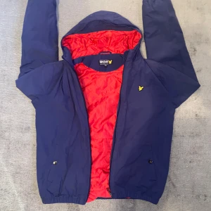 Blå vindjacka från Lyle & Scott - Säljer en snygg blå vindjacka från Lyle & Scott. Den har en cool röd insida och en liten gul logga på bröstet. Jackan har dragkedja och är perfekt för vår och höst. Den är i bra skick och redo för nya äventyr! Strlk xxs eller xs