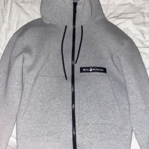 Grå hoodie från Sail Racing - Säljer en stilren grå hoodie från Sail Racing med dragkedja och svart logotyp på bröstet. Insidan är i en kontrasterande gul färg. Perfekt för en casual look med sina långa ärmar och bekväma passform.