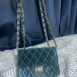 Quiltad axelväska i sammet - Snygg quiltad axelväska i mjuk sammet med en elegant kedjerem. Väskan har en djup grön färg och stängs med ett stilrent spänne i metall. Perfekt för att ge din outfit en lyxig touch.