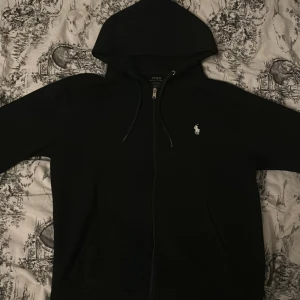 Svart hoodie från Ralph Lauren - Säljer en klassisk svart hoodie från Ralph Lauren med dragkedja och den ikoniska loggan på bröstet. Perfekt för en avslappnad stil. Den har en justerbar huva och fickor framtill.