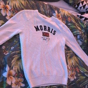 Vit stickad tröja från Morris - Använd 1 gång. Säljer en snygg vit stickad tröja från Morris med text och en brittisk flagga på bröstet. Tröjan har långa ärmar och ribbade muddar vid ärmslut och nederkant. Perfekt för en stilren look!
