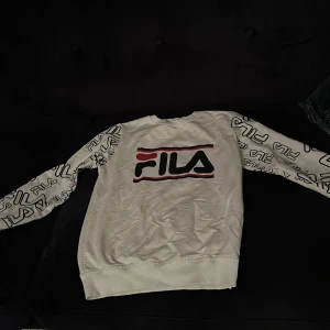 Vit tröja från FILA med logotypmönster - Säljer en vit tröja från FILA med deras ikoniska logotyp i svart och rött på bröstet. Ärmarna har ett mönster av FILA-logotyper. Perfekt för en sportig look eller använda som fin klädnad!