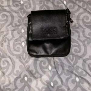 Svart axelväska från Boss - Snygg svart axelväska från Boss i skinn. Väskan har en praktisk dragkedja och en stilren design med Boss-loggan på framsidan. Perfekt för att bära med sig det viktigaste med stil.