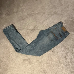 Blå jeans från Nudie Jeans - Snygga blå jeans från Nudie Jeans med en klassisk femficksdesign. De har en knappgylf och är i en slim passform. Perfekta för en avslappnad stil. Storlek 29/30 och modellen är lean Dean.