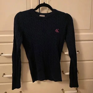 Mörkblå stickad tröja från Calvin Klein - Säljer en snygg mörkblå stickad tröja från Calvin Klein med kabelstickat mönster. Tröjan har långa ärmar och en rund halsringning. 