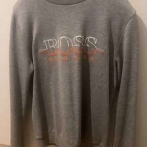Grå sweatshirt från Hugo Boss - Säljer en stilren grå sweatshirt från Hugo Boss med tryckt logga i vitt och orange på framsidan. Tröjan har långa ärmar och rund halsringning. Perfekt för en avslappnad look. Bra skick för det priset du får den för 