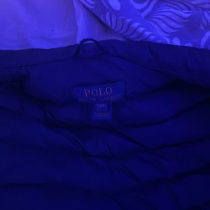 Blå dunjacka från Polo Ralph Lauren - Säljer en snygg blå dunjacka från Polo Ralph Lauren. Jackan har en quiltad design och en dragkedja framtill. Den ikoniska loggan är broderad på bröstet i rött. Perfekt för kyligare dagar!