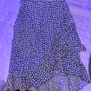 Blommig kjol med volang - Säljer en söt blommig kjol med volangdetaljer. Kjolen har en asymmetrisk design och är perfekt för en somrig look. Den är i en mörk färg med vita blommor som ger en fin kontrast.