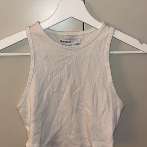 Vit ribbad linnetopp från Bershka - Säljer en stilren vit ribbad linnetopp från Bershka. Perfekt för varma dagar med sin ärmlösa design och bekväma passform. Toppen är enkel att matcha med olika outfits. Hör av dig vid intresse!