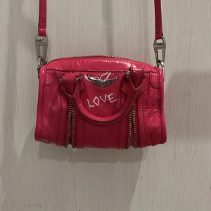 Rosa Zadig axelväska med textdetaljer jätte fin 💕💕 - Snygg rosa axelväska med texten 'LOVE' och 'ZADDICTED' tryckt på framsidan och sidan. Väskan har dubbla handtag och en justerbar axelrem. Den är utrustad med dragkedjor och har en cool, glansig finish.