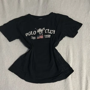 Svart t-shirt från Vinson Polo Club - Snygg svart t-shirt från Vinson Polo Club med tryck av en polospelare och amerikansk flagga på framsidan. Perfekt för en avslappnad stil. T-shirten har korta ärmar och rund halsringning.