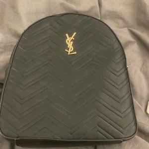 Svart ryggsäck med guldlogga - Snygg svart ryggsäck från YSL med guldlogga på framsidan. Väskan har ett quiltat mönster och dragkedja i guld. Insidan är fodrad i rött tyg och har flera fack för enkel organisering. Perfekt för att bära dina dagliga nödvändigheter med stil.