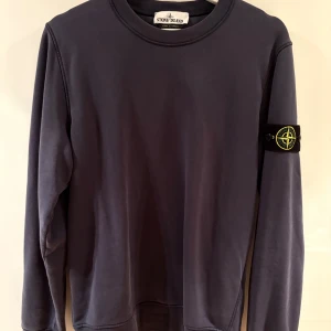 Mörkblå tröja från Stone Island - Säljer en stilren mörkblå tröja från Stone Island med deras ikoniska märke på ärmen. Tröjan har en rund hals och långa ärmar, perfekt för en avslappnad stil. Passar bra till både jeans och chinos.