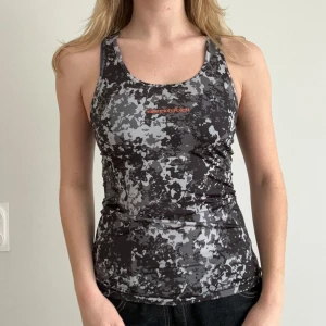 Peak Performance Grå camouflagemönstrat linne - Snygg och sportig grå camouflagemönstrad linnetopp från Peak performance. Perfekt för träning med sin tighta passform och bekväma material. Toppen har ett diskret märkestryck på bröstet och ärmlös design.